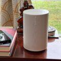 The Sonos Era 100 sits on a table alongside books, a phone, and a lamp near a window.