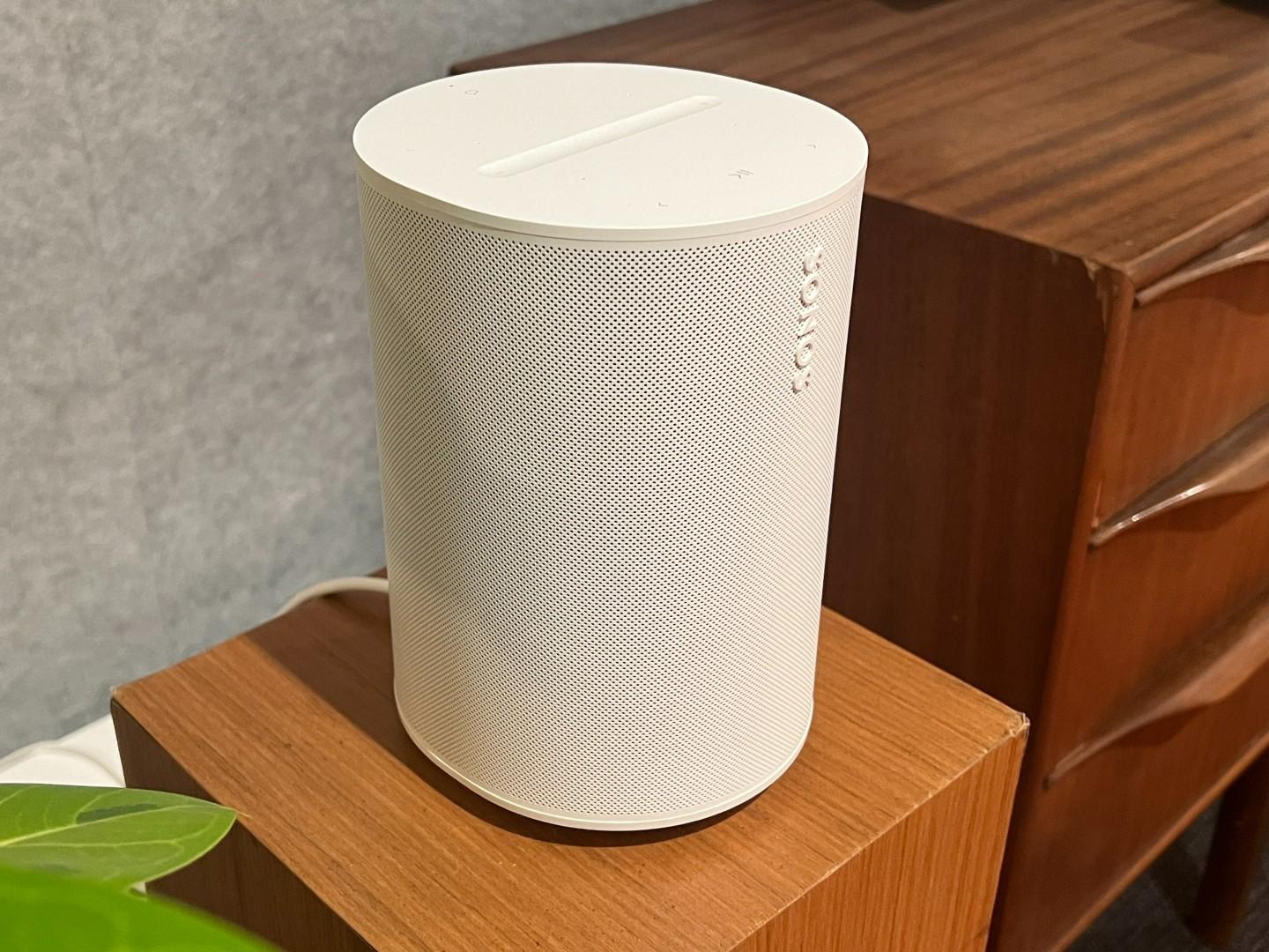 The white Sonos Era 100 smart speaker sits on a light wooden stand in a demo room.