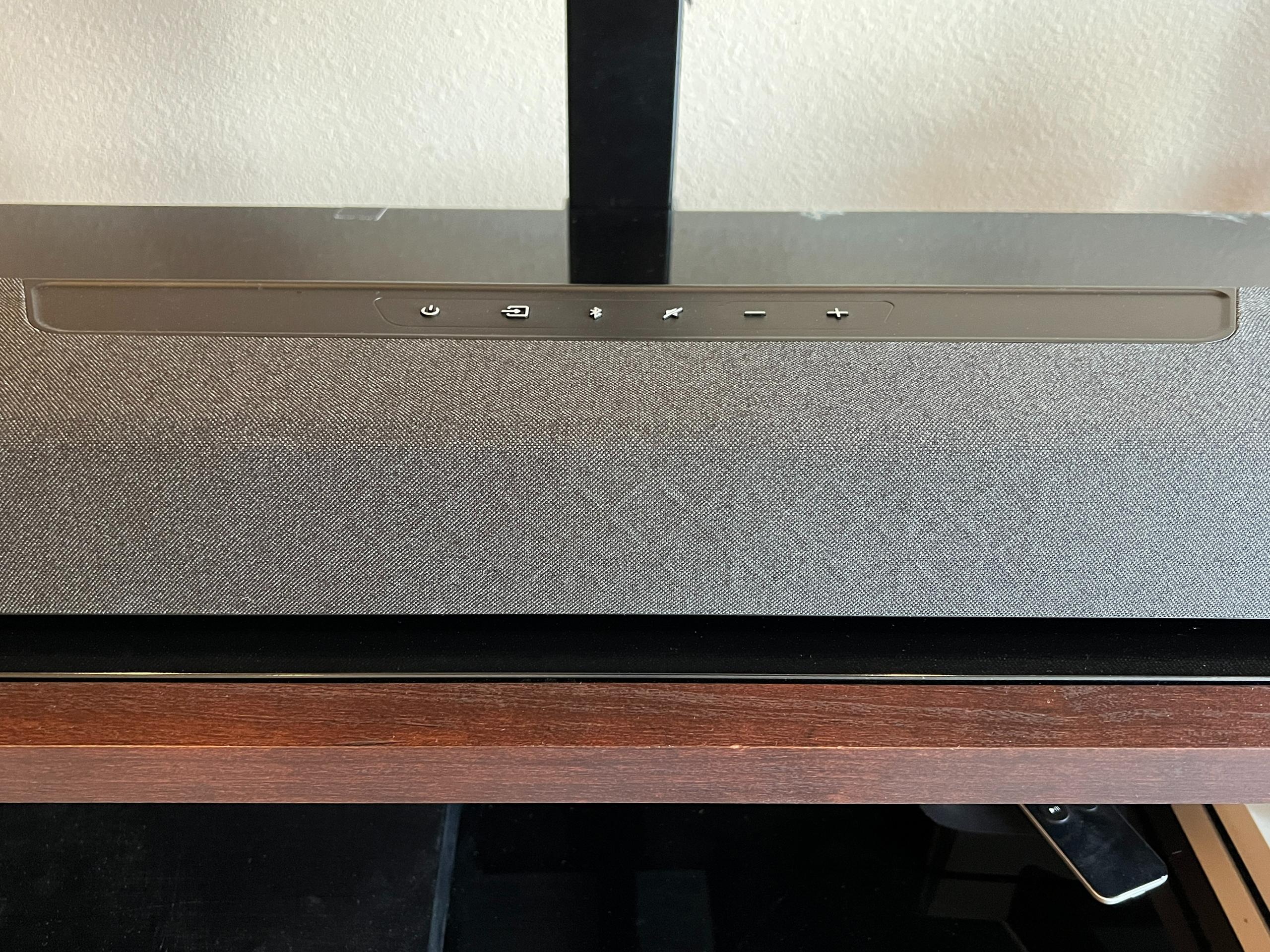 The Polk Magnifi Max AX soundbar controls are shown with the bar atop a black and brown TV console.
